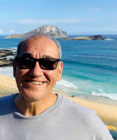 Anthony Ameen Dyer smiling on a shoreline bluff, an island and turquoise surf behind him.